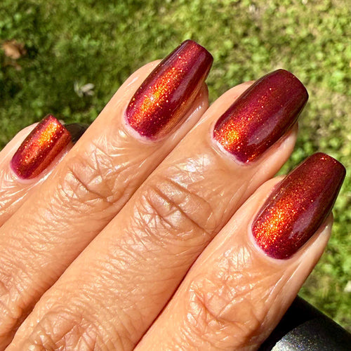 Close-up of a hand with long almond-shaped nails painted in a deep metallic burgundy red with a warm orange-gold shimmer, against a blurred green grass background. A black nail polish bottle is partially visible at the bottom right.